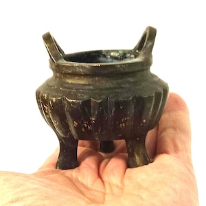 Puede incluir: Un pequeño quemador de incienso antiguo de bronce, con tres patas y dos asas. El recipiente presenta una pátina oscura y envejecida, con signos de desgaste visibles. El interior revela una superficie más oscura, posiblemente oxidada.