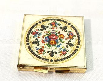 May include: A gold-toned compact mirror with a floral design. The design features a circle of colourful flowers on a white background with a black and gold border.