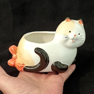May include: A ceramic cat-shaped planter with a white body, brown and orange markings, and black paws and tail. The planter has a smiling face and a hollow interior for plants. The cat is held in a hand against a black background.