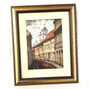 May include: A framed art print featuring a detailed ink and watercolor illustration of a narrow European street scene with historic buildings, red-tiled roofs, and a prominent church tower in the background, enclosed in a textured gold and black frame.