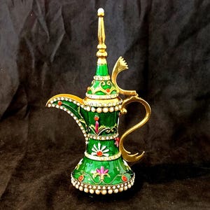 Puede incluir: Una tetera decorativa verde y dorada con intrincados diseños florales y detalles de perlas. La tetera tiene un pico alto y un asa con un remate decorativo.