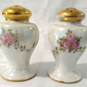 May include: A pair of vintage salt and pepper shakers with a white porcelain base and gold accents. The shakers are decorated with pink roses and green leaves.