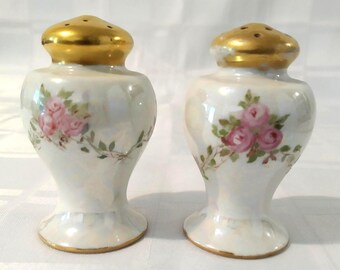 Puede incluir: Un par de saleros y pimenteros vintage con base de porcelana blanca y detalles dorados. Los saleros están decorados con rosas rosas y hojas verdes.