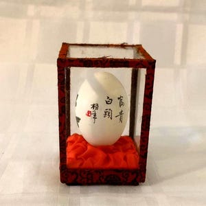 Puede incluir: Un huevo blanco con caligrafía negra en una vitrina de vidrio con un cojín de terciopelo rojo. La caligrafía dice "高青" y "福寿".