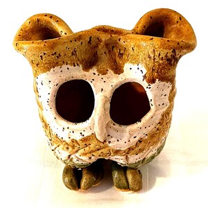 Pode incluir: Um vaso de cerâmica em forma de coruja com esmalte marrom, branco e verde. A coruja tem olhos grandes e um bico pequeno. O vaso tem uma abertura larga e uma base pequena.