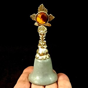May include: A decorative bell with a gold-coloured top and a grey base. The top features a leaf design and a central amber-coloured stone. The bell is held in a hand against a black background.