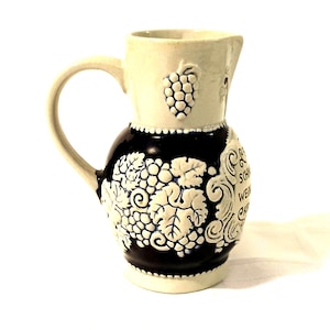 May include: A white and blue ceramic jug with a grape design. The jug has a handle and a spout. The text "SCHWARZWÄLDER WEIN" is on the side of the jug.