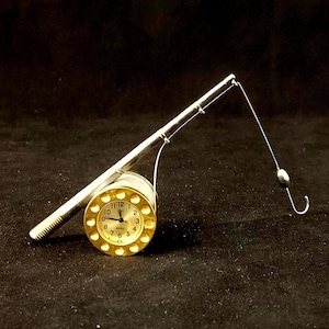 May include: A miniature fishing rod with a gold-coloured clock face. The clock has black numbers and hands, and the rod is silver. The hook and line are also silver, set against a black background.