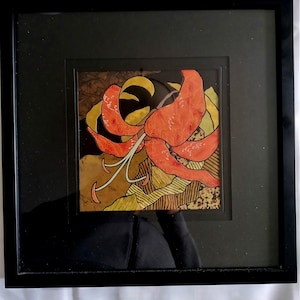 Puede incluir: Una obra de arte enmarcada que presenta una flor de lirio naranja estilizada con detalles intrincados sobre un fondo texturizado. La obra de arte está enmarcada en negro con un borde interior dorado.