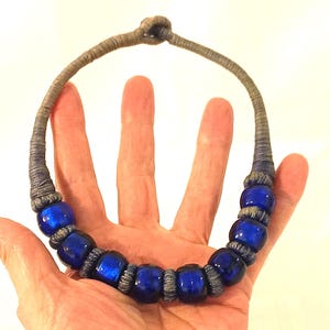 May include: A necklace featuring a series of round, glossy, deep blue beads separated by segments of grey, tightly wrapped thread. The cord is also constructed from the same grey thread, which forms a simple knot closure at the top of the piece.