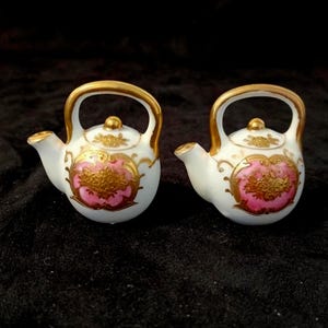 Puede incluir: Dos teteras en miniatura de porcelana blanca con asas y boquillas doradas. Cada tetera está decorada con un panel ovalado rosa central que contiene intrincados patrones florales dorados, con detalles de filigrana dorada en el cuerpo.