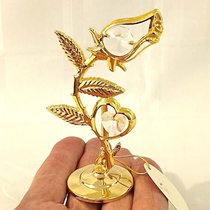 Op de afbeelding: Een goudkleurige metalen roossculptuur met een heldere kristallen rozenknop en een hartvormig kristal. De sculptuur heeft gedetailleerde bladeren en is gemonteerd op een ronde basis. Een klein label hangt aan de basis.