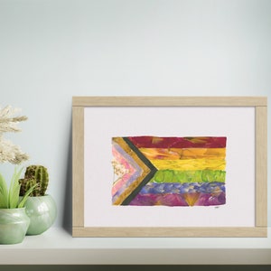 Puede incluir: Una impresión enmarcada de una bandera del orgullo hecha de flores prensadas. La bandera es una variación de la bandera del arcoíris tradicional con una franja negra y marrón añadida en la parte superior.