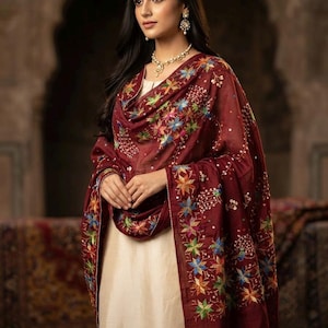 Könnte beinhalten: Ein tiefroter Dupatta mit aufwendiger, mehrfarbiger Blumenstickerei und kleinen reflektierenden Spiegelakzenten, drapiert über einem schlichten cremefarbenen Kleidungsstück. Der Stoff ist transparent und hat detaillierte Nähte an den Rändern.