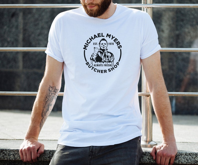 May include: White t-shirt with a black and white graphic of Michael Myers from the Halloween franchise. The graphic is a circle with the text "Michael Myers Est. 1978 Always Fresh Butcher Shop".