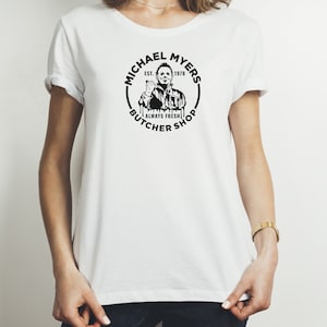 May include: White t-shirt with a black and white graphic of Michael Myers from the Halloween franchise. The graphic is a circle with the text "Michael Myers Est. 1978 Always Fresh Butcher Shop".