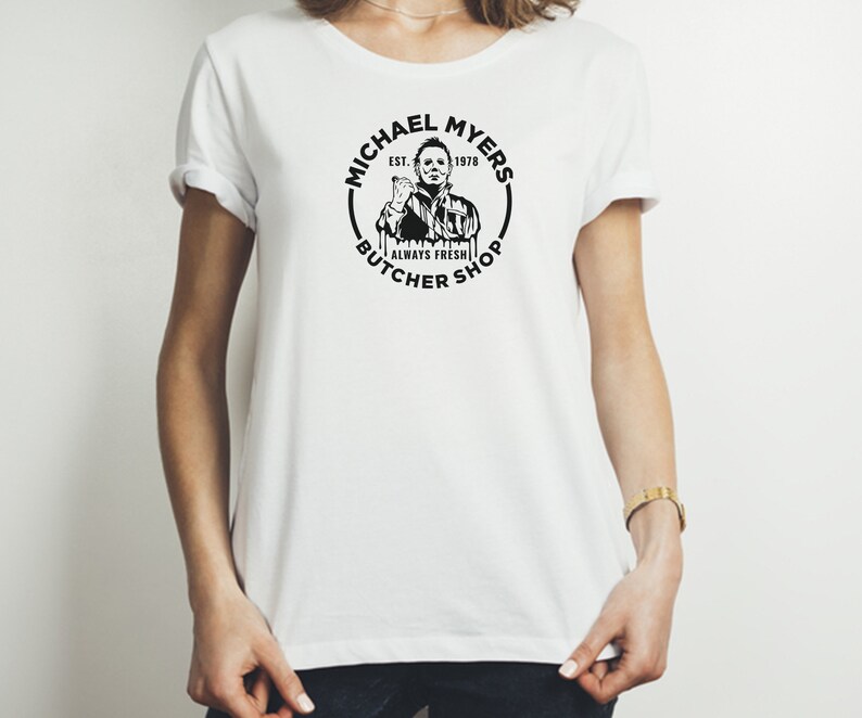 May include: White t-shirt with a black and white graphic of Michael Myers from the Halloween franchise. The graphic is a circle with the text "Michael Myers Est. 1978 Always Fresh Butcher Shop".