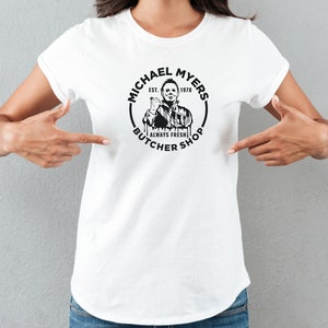 May include: White t-shirt with a black and white graphic of Michael Myers from the movie Halloween. The graphic is a circle with the text "Michael Myers Butcher Shop Est. 1978 Always Fresh" around the image of Michael Myers.