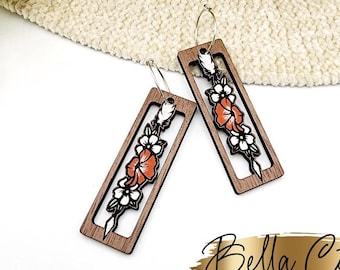 May include: A pair of wooden earrings with a floral design. The earrings are rectangular in shape and have a brown wood finish. The flowers are orange and white and are outlined in black. The earrings are hanging from silver hoops.