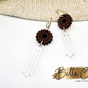 May include: A pair of dangle earrings with a brown and orange wood flower charm and a clear acrylic fringe. The earrings have gold hooks.
