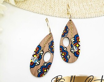 Puede incluir: Un par de pendientes de madera en forma de lágrima con una cadena dorada y una cuenta de cristal transparente. Los pendientes están decorados con un patrón de mandala colorido en tonos de azul, rojo, naranja y amarillo.