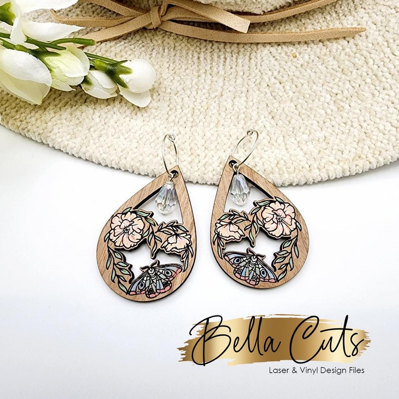 May include: A pair of wooden teardrop-shaped earrings with a floral and butterfly design. The earrings are painted in shades of pink, blue, and green. Each earring features a clear crystal charm.