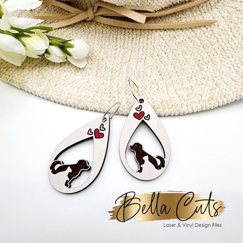 May include: A pair of teardrop-shaped wooden earrings with a silhouette of a dog inside the teardrop. The earrings are decorated with small red hearts.