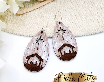 May include: Wooden teardrop earrings featuring a nativity scene design. The earrings have a light wood grain background with a dark brown silhouette of the stable, star, and figures. The earrings are suspended from silver-colored hoops.