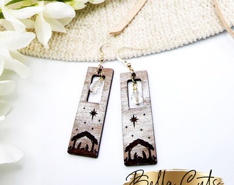 May include: A pair of wooden earrings with a gold-tone finish. The earrings feature a rectangular design with a cutout in the center. The cutout is filled with a clear crystal. The earrings are decorated with a laser-cut design of a nativity scene with a star above the stable. The earrings are hanging from gold-tone hooks.