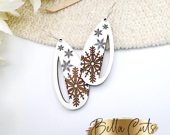 May include: A pair of white wooden earrings with a snowflake design. The snowflakes are cut out of the wood and have a brown stain. The earrings are hanging from silver hooks.