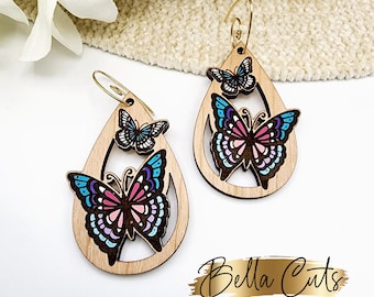 May include: A pair of wooden teardrop-shaped earrings with a layered butterfly design. The butterflies are painted in shades of pink, blue, and purple. The earrings are made of wood and have a gold-coloured metal hook.