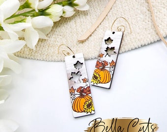 May include: A pair of wooden earrings with a fall theme. The earrings are rectangular and feature a pumpkin, leaves, and a sunflower. The earrings are white with brown accents and have gold hooks.