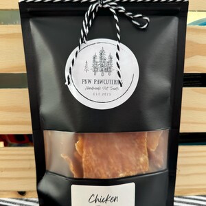 May include: Black bag of chicken jerky with a clear window. The bag has a white label with the words "Chicken Jerky" and a round tag with the text "PNW PAWCUTERIE Handmade Pet Treats EST 2023". The bag is tied with a black and white string.