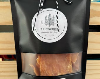 May include: Black bag of chicken jerky with a clear window. The bag has a white label with the words "Chicken Jerky" and a round tag with the text "PNW PAWCUTERIE Handmade Pet Treats EST 2023". The bag is tied with a black and white string.