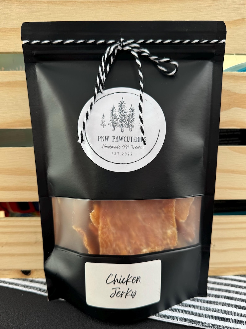 May include: Black bag of chicken jerky with a clear window. The bag has a white label with the words "Chicken Jerky" and a round tag with the text "PNW PAWCUTERIE Handmade Pet Treats EST 2023". The bag is tied with a black and white string.