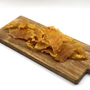 May include: A wooden chopping board displaying several pieces of dried, golden-yellow meat. The meat pieces have a slightly translucent appearance and are arranged in a pile on the board. The background is plain white. The board is light brown.