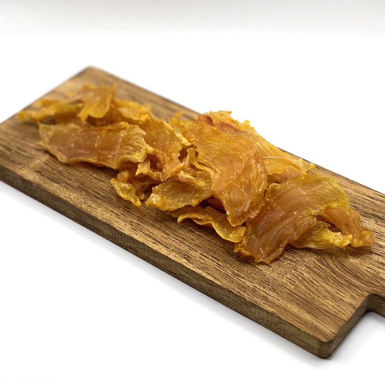 May include: A wooden chopping board displaying several pieces of dried, golden-yellow meat. The meat pieces have a slightly translucent appearance and are arranged in a pile on the board. The background is plain white. The board is light brown.