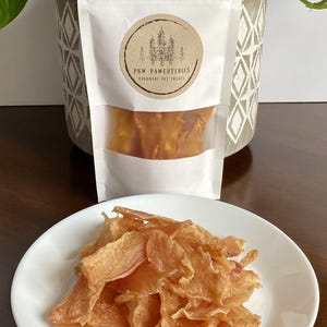 May include: A white bag of handmade pet treats with a clear window, and a round label with a forest design. A white plate holds a pile of light brown, dried pet treats. The label reads "PNW PAWCUTERIES HANDMADE PET TREATS."