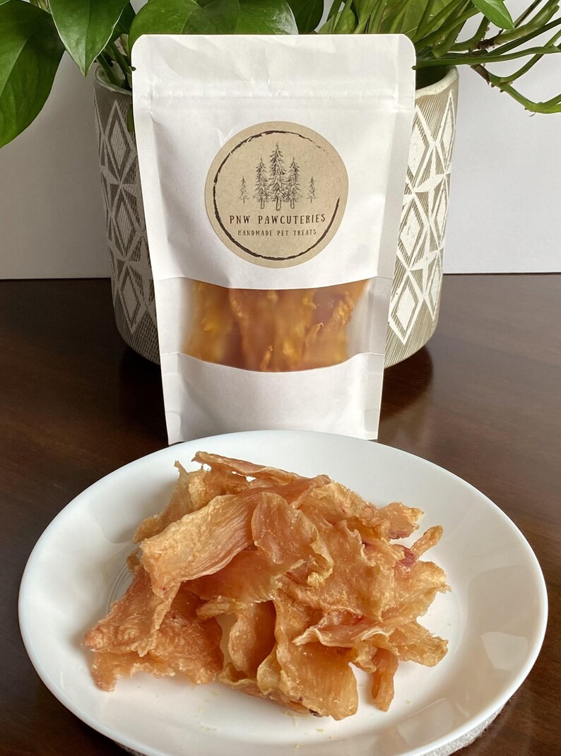 May include: A white bag of handmade pet treats with a clear window, and a round label with a forest design. A white plate holds a pile of light brown, dried pet treats. The label reads "PNW PAWCUTERIES HANDMADE PET TREATS."