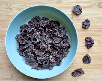 May include: A light blue bowl filled with dark brown, dried food product. Several pieces of the same food are scattered on the light brown wooden surface. The food appears to be dehydrated meat or a similar snack.