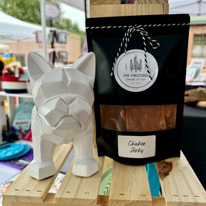 May include: A white geometric dog statue stands beside a black bag of chicken jerky. The bag has a clear window showing the jerky and a label that reads "Chicken Jerky". The bag is tied with a black and white string and has a round label with a tree design.