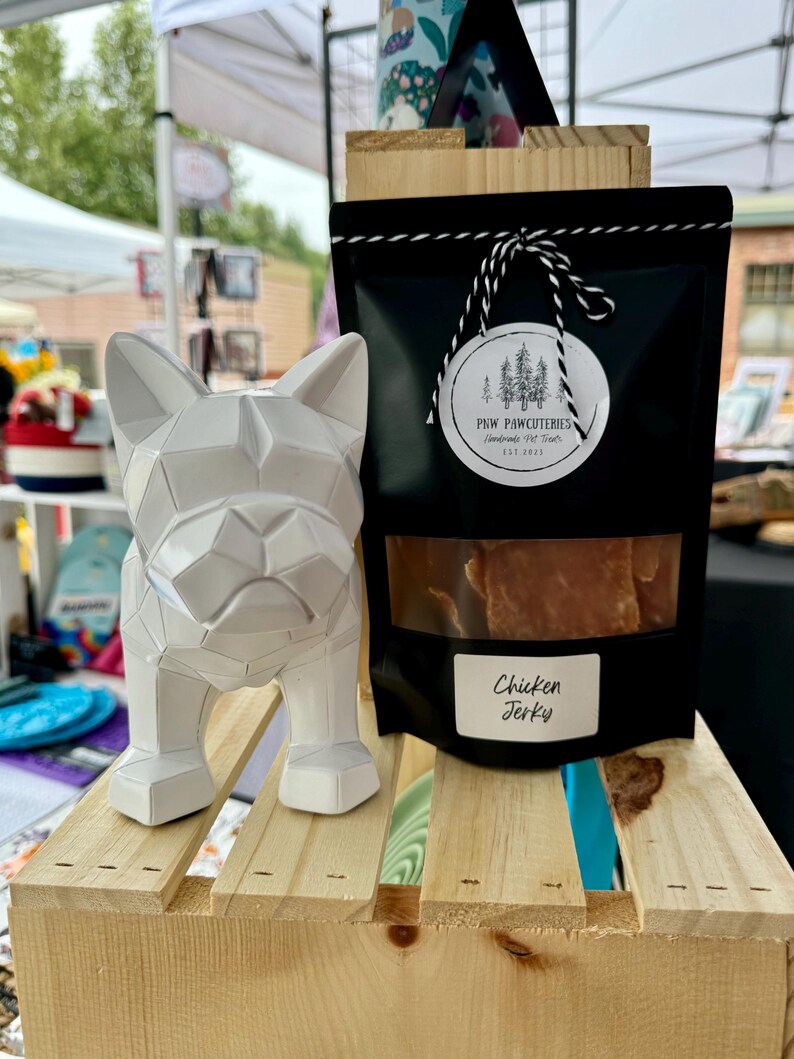 May include: A white geometric dog statue stands beside a black bag of chicken jerky. The bag has a clear window showing the jerky and a label that reads "Chicken Jerky". The bag is tied with a black and white string and has a round label with a tree design.