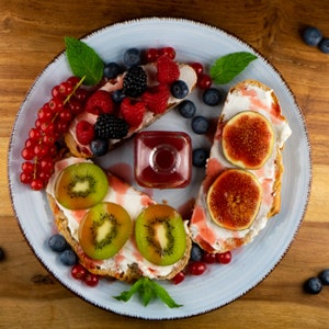 May include: A light blue plate with assorted fruit and toast. The toast is topped with cream cheese, kiwi, figs, and berries. A small square glass bottle of red liquid sits in the center. Fresh mint leaves are scattered around the plate.