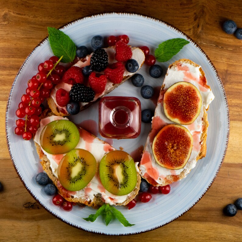 May include: A light blue plate with assorted fruit and toast. The toast is topped with cream cheese, kiwi, figs, and berries. A small square glass bottle of red liquid sits in the center. Fresh mint leaves are scattered around the plate.
