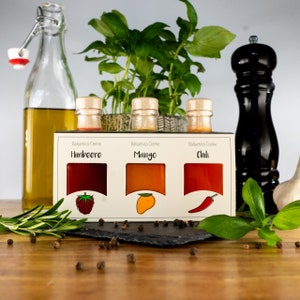 May include: A display of gourmet food items. A clear glass bottle with olive oil, a box of balsamic cream with raspberry, mango, and chili flavors, a black pepper grinder, and fresh herbs are arranged on a wooden surface.