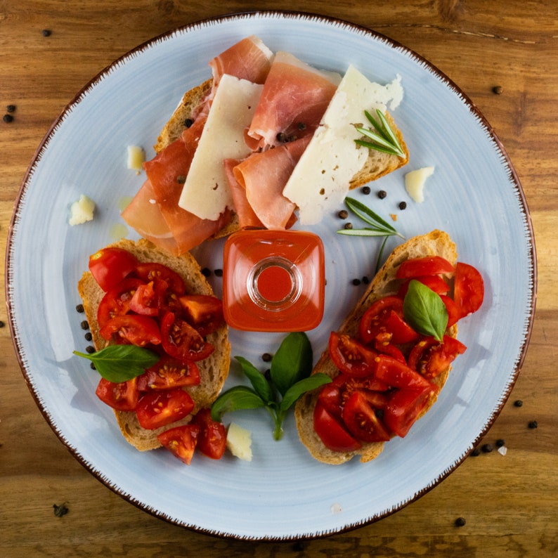 May include: A light blue plate with a variety of Italian appetizers. Bruschetta with diced tomatoes and basil, prosciutto, and cheese slices are arranged around a small square bottle of red sauce. Black peppercorns and rosemary sprigs complete the presentation.