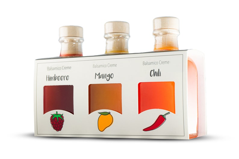 May include: Three glass bottles of balsamic cream with wooden stoppers, presented in a white box. The flavors are raspberry, mango, and chili, each with a corresponding fruit or pepper illustration. The labels read "Balsamico Creme".