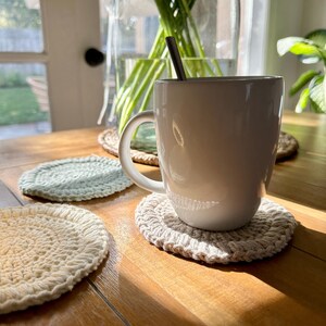 May include: A white ceramic mug with a metal spoon resting on the rim, sitting on a beige crocheted coaster. Two other crocheted coasters, one green and one beige, are visible on a wooden table.