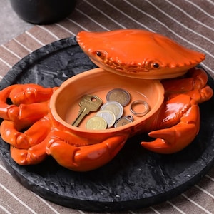 Könnte beinhalten: Orangefarbene Krabben-Schmuckschale auf einem schwarzen Untergrund in Marmoroptik. Die Schale enthält Schlüssel, Münzen und einen Ring. Die obere Schale des Krebses hebt sich, um den Stauraum freizulegen. Sie liegt auf einer gestreiften braun-weißen Oberfläche.