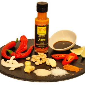 Könnte beinhalten: Ein schwarzer Schieferteller mit Zutaten für die Zubereitung von Satay, darunter rote Chilischoten, Knoblauch, Ingwer, Erdnüsse, Kokosflocken, Limettenspalten, Salz, Zucker und eine Flasche Chillisauce mit der Aufschrift "CHILMA Gourmet Chili Sauce Satay 100ml Scharfe 3 von 7".